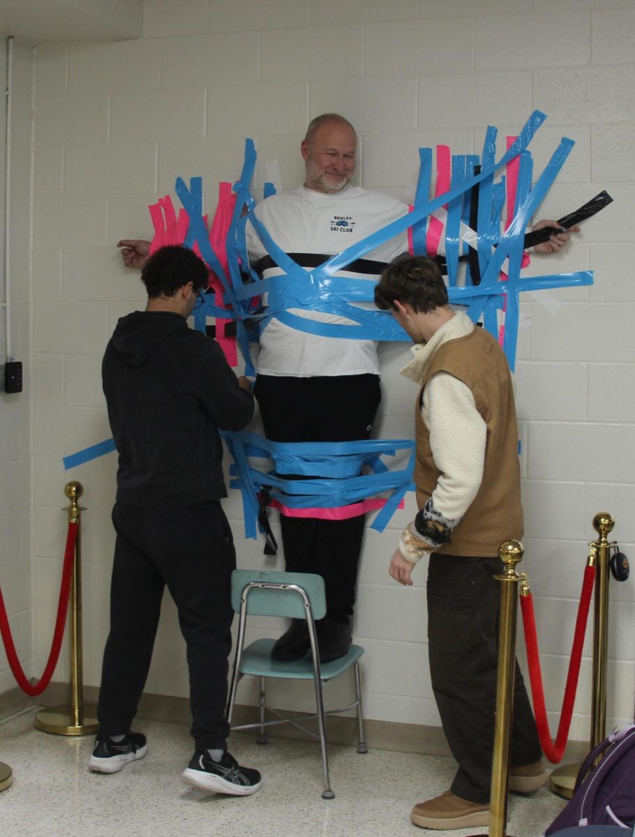 Sophomore Orion Abner and senior Wilson Klingelhofer tape Principal Jason Caudill to the cafeteria wall to raise money for DARN.